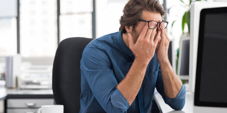 Portrait of an upset businessman at desk in office. Businessman being depressed by working in office. Young stressed business man feeling strain in eyes after working for long hours on computer.